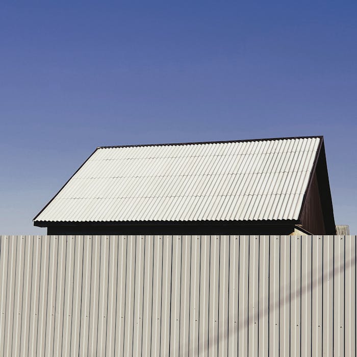 gallery-03 A simple structure with a metal roof and fence under a clear blue sky, showcasing minimalist architecture.
